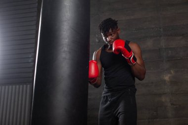 Genç, güçlü, kırmızı eldivenli Afrikalı Amerikalı boksör kum torbasına vuruyor, kaslı atlet trenleri koyu renk spor salonunda yumrukluyor ve siyah arka planda spor yapıyor.
