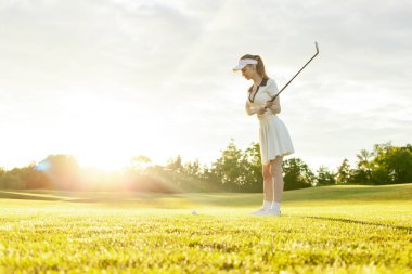 Spor üniformalı genç ve çekici bir kız gün batımında çimlerle golf oynuyor. Golf sopalı bir kadın açık havada spor yapıyor.