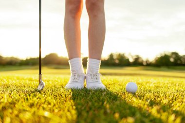 Genç kız golf sahasında golf oynuyor ve baloda salıncaklar, yakın plan beyaz ayaklı golf ayakkabıları ile çimenlerin ve günbatımının arka planında.