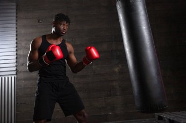 Genç, güçlü, kırmızı eldivenli Afrikalı Amerikalı boksör kum torbasına vuruyor, kaslı atlet trenleri koyu renk spor salonunda yumrukluyor ve siyah arka planda spor yapıyor.