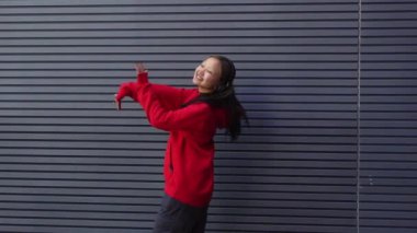 Kırmızı kapüşonlu genç kız kulaklıkla müzik dinliyor ve sokakta dans ediyor, enerjik kadın hip hop dansçısı şehrin arka planında duvarın arkasında dans ediyor.