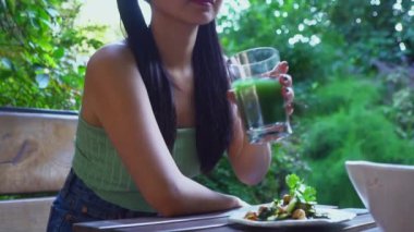 Genç Asyalı kadın, yeşil bardaktan içer ve sokak kafesinde gülümser Koreli kadın, parktaki caddedeki restoranda sebzeli smoothie içer.