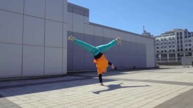 Genç enerjik erkek dansçı açık havada break dans yapıyor ve şehrin arka planında duvara karşı el üstünde zıplıyor, erkek akrobat ekstrem numaralar yapıyor ve hip-hop yapıyor.