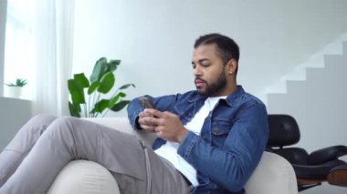 Üzgün Afrikalı Amerikalı adam akıllı telefon kullanıyor ve evde online olarak kaybediyor, adam sandalyede oturuyor cep telefonunda daktilo yazıyor ve başarısızlıktan facepalm gösteriyor