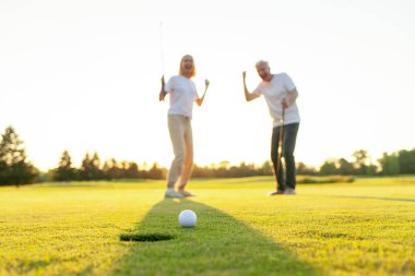 Yaşlı adam ve kadın golfün galibiyetini kutluyor ve el kaldırarak bağırıyor, yaşlı çift gün batımında golf oynuyor ve golf ekipmanlarıyla başarıyı kutluyor.