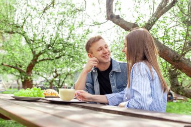 Kot elbiseli genç bir çift ağaçtan bir masanın bahçesinde oturuyor ve yaz mevsiminde konuşuyorlar. Erkek ve kız parkta konuşup flört ediyorlar.