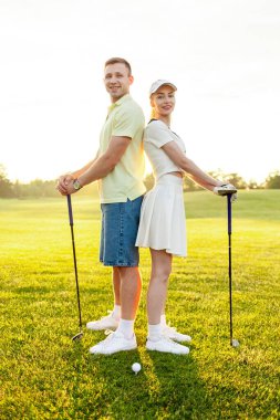 Üniformalı genç bir çift gün batımında çimenlikte golf oynuyor, eğitmenli bir kadın golf antrenmanı yapıyor ve aktif olarak dinleniyor.