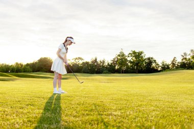 Spor üniformalı genç ve çekici bir kız gün batımında çimlerle golf oynuyor. Golf sopalı bir kadın açık havada spor yapıyor.