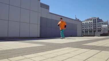 Genç enerjik erkek dansçı açık havada break dans yapıyor ve şehrin arka planında duvarın karşısında ayakta duruyor erkek akrobat ekstrem numaralar yapıyor ve hip-hop dansı yapıyor.