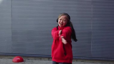 Kırmızı kapüşonlu genç kız kulaklıkla müzik dinliyor ve sokakta dans ediyor, enerjik kadın hip hop dansçısı şehrin arka planında duvarın arkasında dans ediyor.
