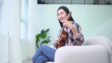 Genç Asyalı kadın ukulele çalıyor, evinde oturuyor ve gülümsüyor, Koreli bayan müzisyen beyaz odada akustik müzik aleti çalıyor.