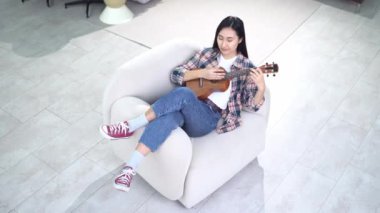 Genç Asyalı bir kadın evinde rahat bir sandalyede uzanıp ukulele çalıyor ve hayal kuruyor, bayan müzisyen akustik gitar çalıyor ve hayal ediyor.