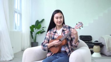 Genç Asyalı kadın ukulele çalıyor, evinde oturuyor ve gülümsüyor, Koreli bayan müzisyen beyaz odada akustik müzik aleti çalıyor.