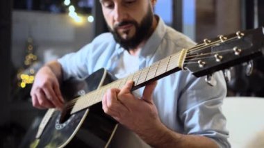Gömlekli sakallı adam evde siyah akustik gitar çalıyor, müzisyen içeride müzik aleti kullanıyor ve şarkı söylüyor, yakın plan.