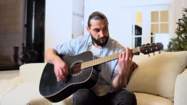 Yılbaşı için evde akustik gitar çalan sakallı bir adam Noel ağacının yanındaki koltukta oturan ve yılbaşı için müzik aleti kullanan bir adam.