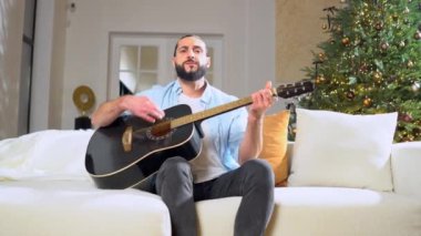 sakallı adam akustik gitar çalıyor ve noel için evde şarkı söylüyor, noel ağacının yanındaki koltukta oturuyor ve yeni yıl için müzik aleti kullanıyor.