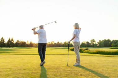 Yaşlı çift golf sahasında golf oynuyor ve gün batımında sohbet ediyor. Yaşlı erkek ve kadın dışarıda dinleniyor ve gülüyor. Emekliler tatilde.