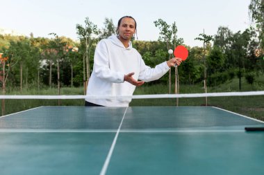 Parkta açık havada masa tenisi oynayan genç Afrikalı Amerikalı bir adam, bir adam pinpon raketiyle topa vuruyor.