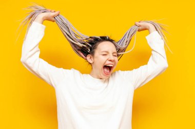 Renkli rastalı genç ve sinirli bir kız sarı arka planda saçlarını tutarak çığlık atıyor. Benzersiz saç stili ve saç örgüleri olan bir kadın stresten panik yapıyor.