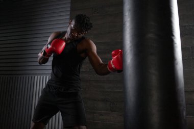 Genç, güçlü, kırmızı eldivenli Afrikalı Amerikalı boksör kum torbasına vuruyor, kaslı atlet trenleri koyu renk spor salonunda yumrukluyor ve siyah arka planda spor yapıyor.