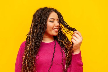 Saçları örgülü neşeli, tombul Afro-Amerikan kadın saçlarını tutuyor ve sarı yalıtılmış arka planda gülümsüyor. Rasta saçlı, iri yarı, iri yarı bir kız.
