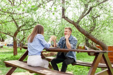 Kot elbiseli genç bir çift, tahta bir masada bahçede oturuyor ve yazın bir randevuda kahve içiyor, kız ve erkek açık hava kafede kahvaltı yapıyor ve sohbet ediyorlar.