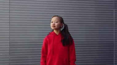 Kırmızı kapüşonlu neşeli genç Asyalı kız şehir caddesinde duruyor ve gülümsüyor. Koreli kadın şehir duvarının yanında duruyor ve saçını dışarıda düzleştiriyor.