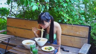 Genç, neşeli Asyalı bir kadın açık kafede çubuklarla ramen yiyor. Parktaki bir restoranda erişte yiyor.