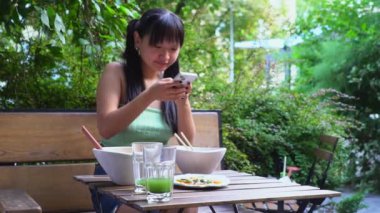 Genç, neşeli Asyalı kadın açık hava kafede akıllı telefondan yemek fotoğrafı çekiyor, kadın blogcu, parktaki restoranda öğle yemeği yiyor ve cep telefonuyla fast food fotoğrafları çekiyor.