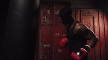 Afrika kökenli Amerikalı genç boksör. Koyu renk spor salonunda antrenman yapıyor. Eldivenlerle kum torbasına vuruyor.