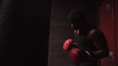 Afrika kökenli Amerikalı genç boksör. Koyu renk spor salonunda antrenman yapıyor. Eldivenlerle kum torbasına vuruyor.