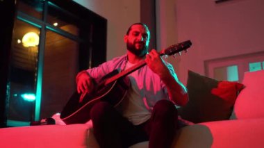 Evde akustik gitar çalan sakallı bir adam neon ışıkta, müzisyen gece müzik enstrümanıyla şarkı söylüyor.