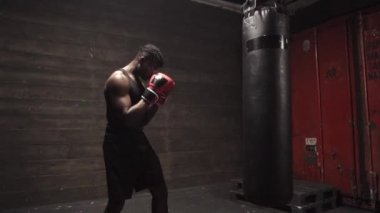 Afrika kökenli Amerikalı genç boksör. Koyu renk spor salonunda antrenman yapıyor. Eldivenlerle kum torbasına vuruyor.