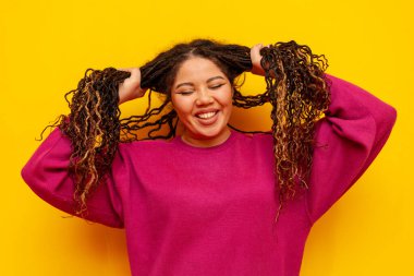 Saçları örgülü neşeli, tombul Afro-Amerikan kadın saçlarını tutuyor ve sarı yalıtılmış arka planda gülümsüyor. Rasta saçlı, iri yarı, iri yarı bir kız.