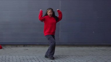 Kırmızı kapüşonlu genç kız kulaklıkla müzik dinliyor ve sokakta dans ediyor, enerjik kadın hip hop dansçısı şehrin arka planında duvarın arkasında dans ediyor.