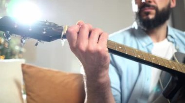 Gömlekli sakallı adam evde siyah akustik gitar çalıyor, müzisyen içeride müzik aleti kullanıyor ve şarkı söylüyor, yakın plan.