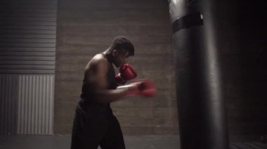 Afrika kökenli Amerikalı genç boksör. Koyu renk spor salonunda antrenman yapıyor. Eldivenlerle kum torbasına vuruyor.