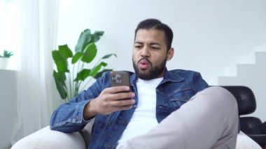 Şok olmuş Afro-Amerikan adam evde akıllı telefon kullanıyor ve şaşırmış, heyecanlı adam telefona bakıyor ve şaşırmış bir şekilde daktilo yazıyor.