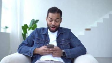 Şok olmuş Afro-Amerikan adam evde akıllı telefon kullanıyor ve şaşırmış, heyecanlı adam telefona bakıyor ve şaşırmış bir şekilde daktilo yazıyor.