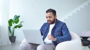 Heyecanlı Afrikalı Amerikalı adam dizüstü bilgisayar kullanıyor ve evinde online olarak kazanıyor, kazanan adam başarıyı kutluyor ve koltukta otururken iyi şanslar.