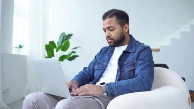 Ciddi bir Afro-Amerikan erkeği dizüstü bilgisayar kullanıyor ve evde internetten yazıyor. Adam çalışıyor ve bir sandalyede otururken bilgisayara bakıyor.
