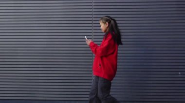 Kırmızı kapüşonlu neşeli Asyalı kız şehrin arka planında akıllı telefon kamerasıyla fotoğraf çekiyor. Koreli kadın cep telefonuyla şehri filme çekiyor.