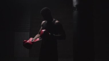 Afro-Amerikan atletik bir adam karanlık bir odada kırmızı boks eldivenleri giyiyor, güçlü bir boksör eğitime ve dövüşmeye hazırlanıyor.
