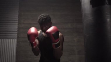 Afrika kökenli Amerikalı genç boksör, karanlık spor salonunda antrenman yapıyor ve kameraya eldivenle vuruyor, güçlü bir adam spor yapıyor ve dayanıklılık eğitimi alıyor.