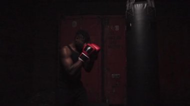 Afrika kökenli Amerikalı genç boksör. Koyu renk spor salonunda antrenman yapıyor. Eldivenlerle kum torbasına vuruyor.