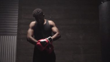 Afro-Amerikan atletik bir adam karanlık bir odada kırmızı boks eldivenleri giyiyor, güçlü bir boksör eğitime ve dövüşmeye hazırlanıyor.