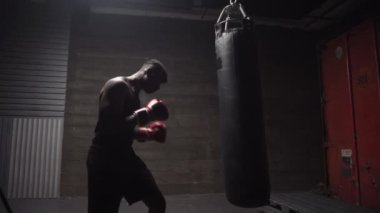 Afrika kökenli Amerikalı genç boksör. Koyu renk spor salonunda antrenman yapıyor. Eldivenlerle kum torbasına vuruyor.