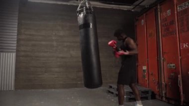 Afrika kökenli Amerikalı genç boksör. Koyu renk spor salonunda antrenman yapıyor. Eldivenlerle kum torbasına vuruyor.