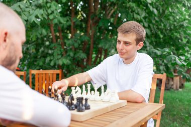 İki genç adam ahşap masada oturuyor ve parkta satranç oynuyorlar. Erkek öğrenciler masa oyununda yarışıyorlar ve strateji planlıyorlar.