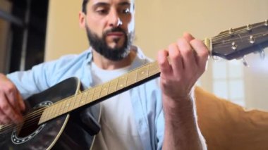 Gömlekli sakallı adam evde siyah akustik gitar çalıyor, müzisyen içeride müzik aleti kullanıyor ve şarkı söylüyor, yakın plan.